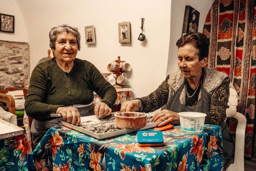 Two older women sit at a table as they sort and clean mastic resin drops from soil and debris.