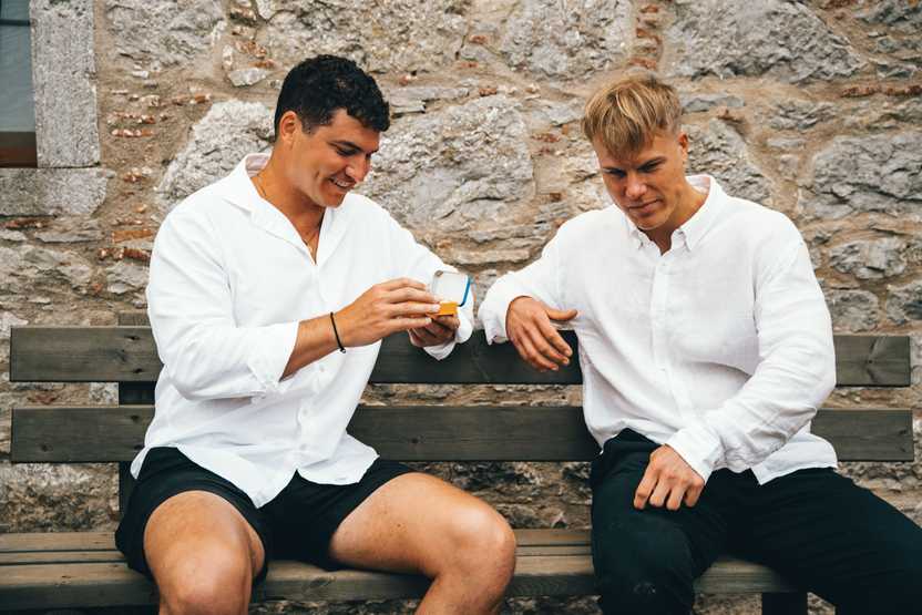 Two young men in white linen shirts sitting on a bench chewing mastic gum, one of them is holding a tin of Greco Gum