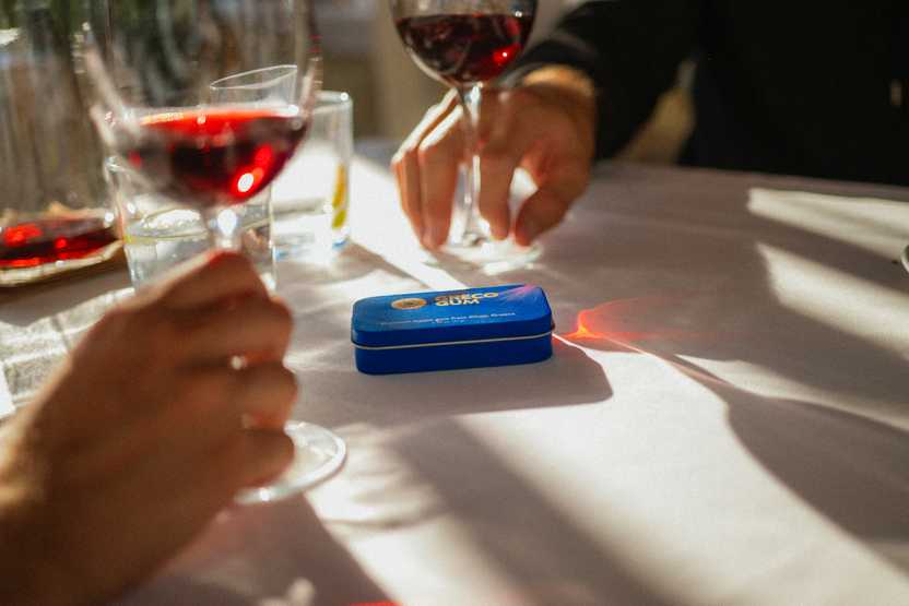 Greco Gum tin of mastic droplets on a white-cloth dinner table, you can also see the hands of two different people each holding a glass of red wine.